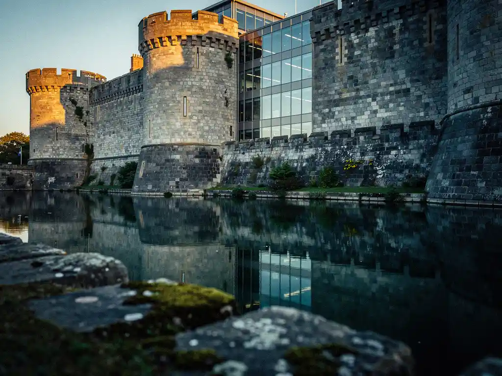 Ancient stone castle moat at golden hour reflecting a modern glass office building, slate grey fortress walls bathed in warm amber light.