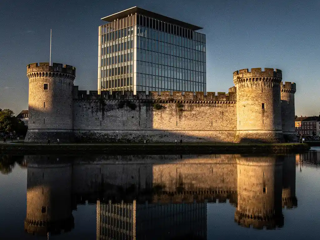 Stone castle moat reflecting still water, with a modern glass corporate building rising seamlessly behind medieval walls in golden afternoon light.
