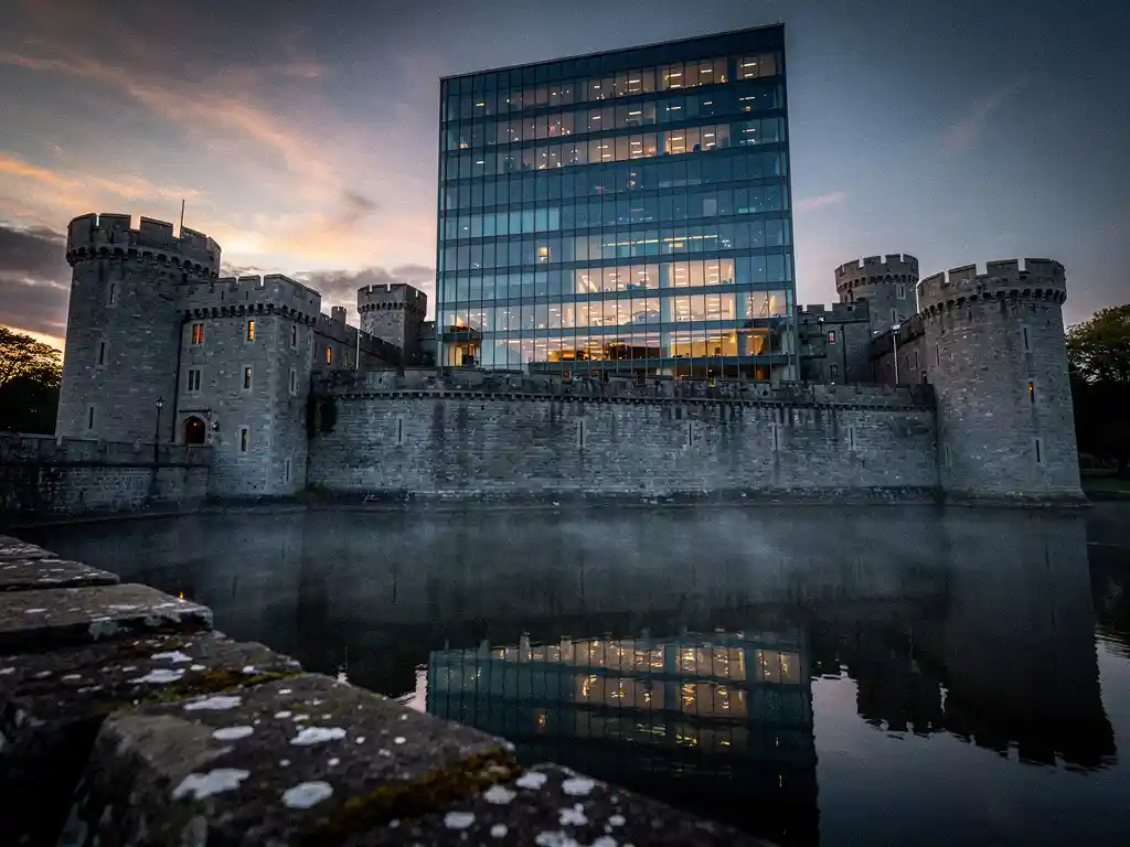 Medieval stone castle with moat reflecting a modern glass office tower rising from its walls at dusk, blending historic and contemporary architecture.